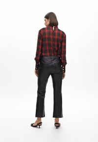 Blusa trasparente a quadri rossi e neri con dettaglio a chiave sulla schiena, abbinata a pantaloni di pelle neri a vita alta. L'outfit presenta dettagli con bottoni dorati.