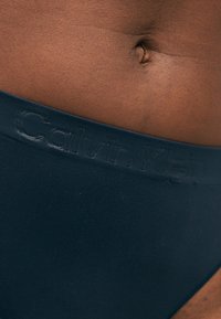 Κοντινό πλάνο στη μέση ενός ατόμου με σκούρο δέρμα, μαύρη μέση με ανάγλυφο κείμενο "Calvin Klein" και εμφανή ομφαλό.