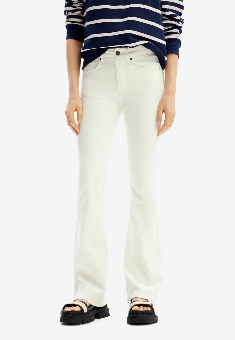 Personne portant un jean blanc à taille haute évasé, une chemise à rayures bleu marine et blanche à manches longues, et des sandales à plateforme noires avec des bretelles beiges.