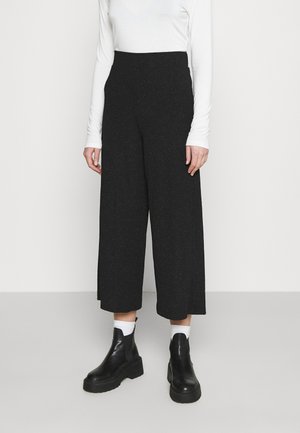 Personne portant un pantalon noir large taille haute, un haut blanc à manches longues, des chaussettes blanches et des bottines plateformes noires, debout devant un fond uni.