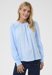 Blouse à manches longues bleu clair avec un motif de pois texturé, encolure froncée et poignets élastiqués, assortie à un jean foncé.