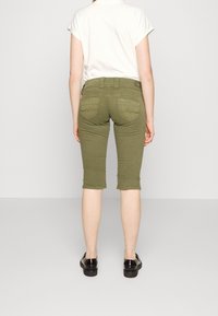 Osoba stojící s zády k fotoaparátu, oblečená v olivově zelených 3/4 kalhotách, bílé košili s krátkým rukávem a černých mokasínech na jednobarevném pozadí.