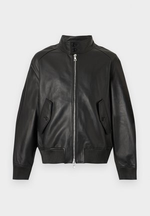 Homme debout portant une veste en cuir noire par-dessus une chemise blanche, un pantalon beige et des chaussures en cuir noires sur fond blanc.