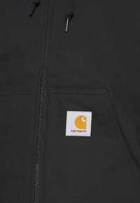 Black zip-up jacket with a textured fabric. Features a square white label with a yellow logo on the chest pocket. Adjustable drawstrings.
