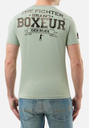 Homme portant un t-shirt vert clair avec l'inscription « THE FIGHTER BRAND BOXEUR DES RUES » et une silhouette de boxe imprimée au dos, assorti à un jean bleu.