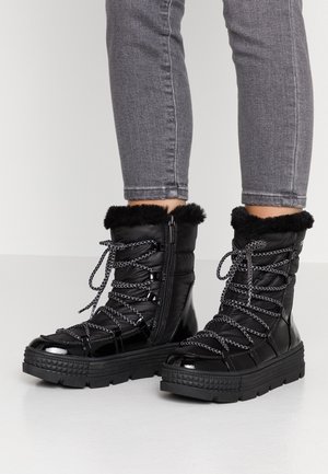 Sorte vinterstøvler til midt på læggen med pelskant, mønstrede snørebånd, sidelukning med lynlås, båret med grå tætsiddende jeans.