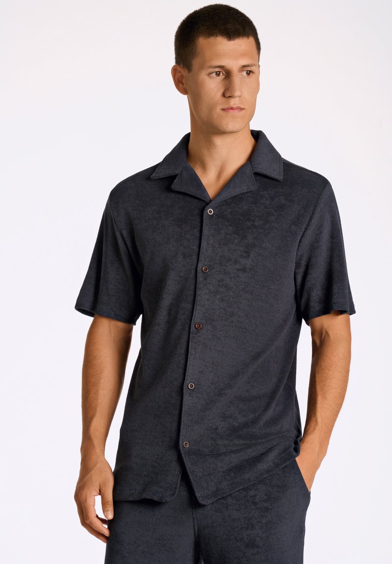 Camicia a maniche corte, con bottoni, di colore grigio scuro, realizzata in tessuto morbido con una superficie testurizzata e colletto classico. Presenta bottoni marroni.