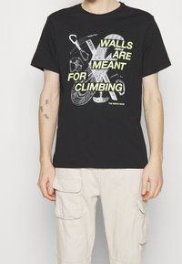 Černé bavlněné tričko s grafickým designem, který zobrazuje horolezecké nástroje a text "ZDI JSOU URČENY K LEZENÍ" v jasně žluté barvě.