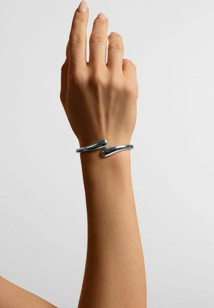 Silverfärgat metalarmband med en mjuk, böjd design. Armbandet har två tydliga öglor med en polerad yta, vilar på en hand.