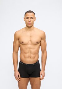 Γυμνασμένος άνδρας που στέκεται όρθιος, βλέποντας μπροστά, φορώντας μαύρα εσώρουχα Tommy Hilfiger με λογότυπο στη μέση, σε απλό λευκό φόντο.