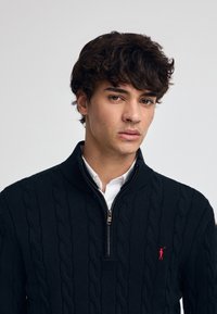 Pull en maille torsadée bleu marine avec col zippé mi-haut et un petit logo rouge sur la poitrine. Le matériau a une texture douce.