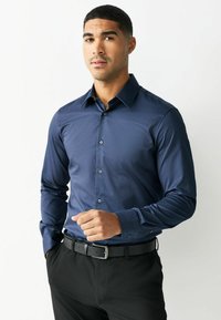 Man die een lange-mouwen, marineblauw overhemd met knopen draagt, ingestopt in een zwarte broek met een zwarte riem, staand tegen een witte achtergrond.