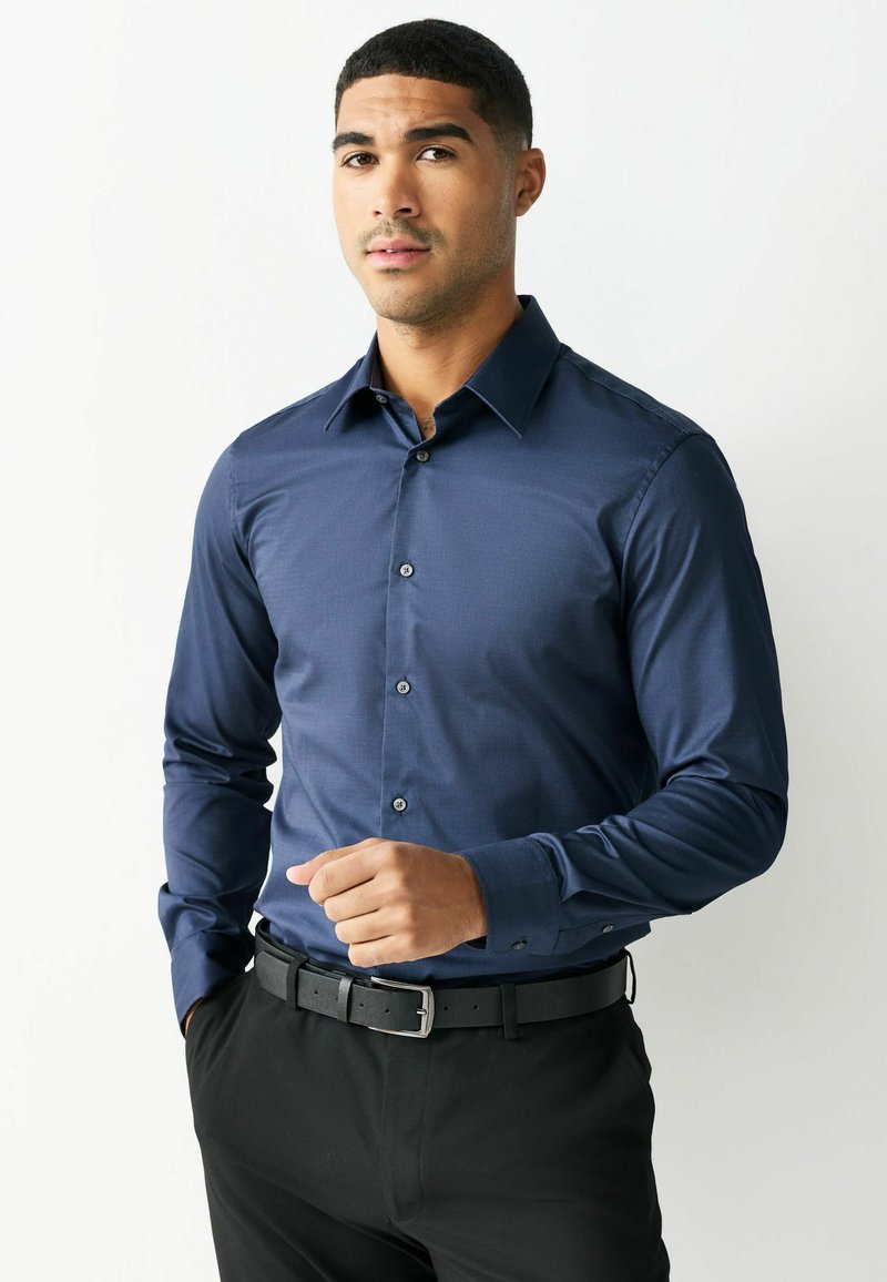 Man die een lange-mouwen, marineblauw overhemd met knopen draagt, ingestopt in een zwarte broek met een zwarte riem, staand tegen een witte achtergrond.