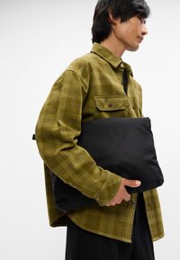 Sac rembourré noir tenu contre une personne portant une chemise à carreaux verts. La chemise a des poignets boutonnés et deux poches poitrine.