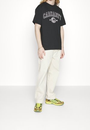 Person iført sort Carhartt T-shirt, hvide bukser og grøn-gule sneakers, stående mod en ensfarvet hvid baggrund.