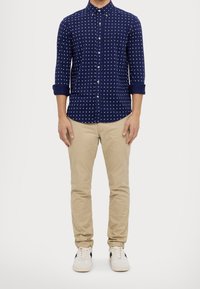 Námornická košile na knoflíky s bílým čtvercovým vzorem, vyhrnut é rukávy, spojená s béžovými chinos kalhotami a bílými teniskami s černými detaily.