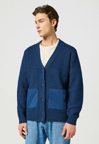 Granatowy sweter z dzianiny o fakturowanym wzorze, z dwiema solidnymi granatowymi kieszeniami na łatki, dekoltem w kształcie V i zapięciem na guziki. Nosi się go na jasną bluzkę.