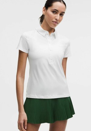 Femeie purtând un tricou polo alb cu mâneci scurte și o fustă plisată verde închis pe un fundal simplu.