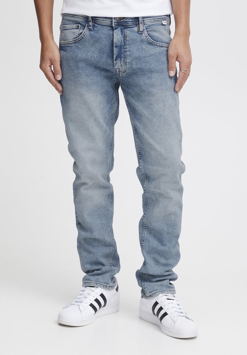 Jeans de mezclilla de color azul claro con corte recto, cinco bolsillos y una textura desgastada. Combinados con zapatillas blancas con rayas negras.