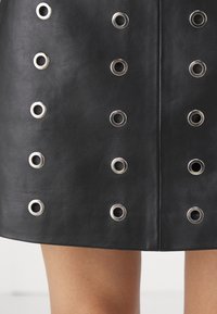 Jupe en cuir noir avec des œillets métalliques argentés disposés en rangées verticales au-dessus de jambes nues.