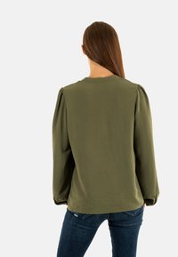Blusa de manga larga verde olivo con un acabado texturizado, cuello redondo y detalle fruncido en el hombro, combinada con jeans de color azul oscuro.