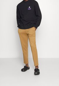Schwarzes Sweatshirt mit lila Logo, beigefarbene tapered Hose und schwarze Clogs. Das Outfit kombiniert legere und zeitgemäße Designelemente.