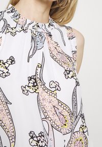 Femme portant un chemisier blanc sans manches à col montant avec un grand motif floral paisley pastel en rose doux, jaune et noir.