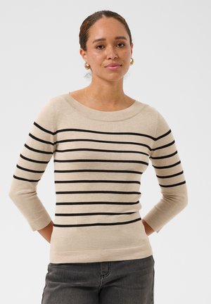 Femme à la peau marron clair et aux cheveux tressés, portant un pull beige à rayures horizontales noires et un jean gris foncé, debout les mains dans le dos.