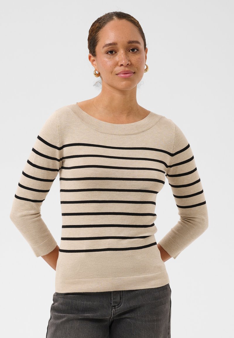 Femme à la peau marron clair et aux cheveux tressés, portant un pull beige à rayures horizontales noires et un jean gris foncé, debout les mains dans le dos.