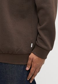 Brązowa bluza z miękkiego materiału z prążkowanymi mankietami i pasem. Z boku znajduje się mała naszywka z logo. Tekstura wydaje się gładka i wygodna.
