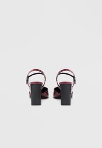 Bordeauxrode sandalen met hoge hakken, glanzende afwerking, gesloten neus en enkelriem. Voorzien van een stevige zwarte hak en een gladde textuur.