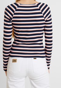 Langarm gestreiftes Oberteil in Marineblau, Pink und Hellblau. Besitzt ein tailliertes Design und eine gerippte Textur. Getragen mit weißen Jeans und einem sichtbaren Logo-Tag.