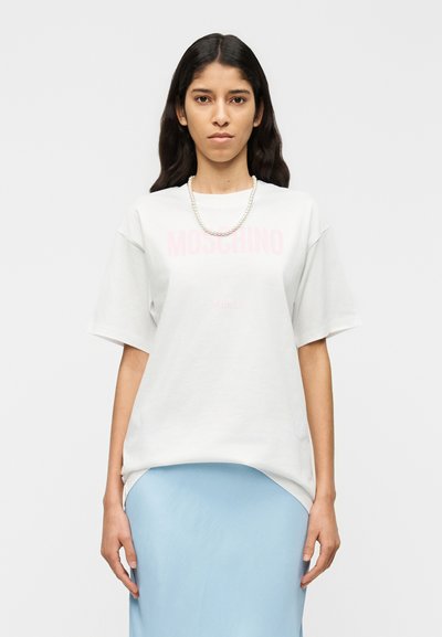 Λευκό oversized t-shirt με ροζ γραφικό "MOSCHINO". Κοντά μανίκια, στρογγυλή λαιμόκοψη. Φοριέται με φούστα από σατέν σε ανοιχτό μπλε χρώμα.