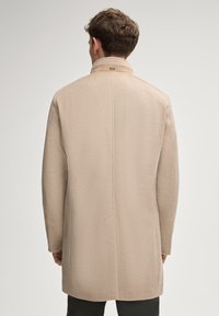 Beige wollen jas met een gestructureerd ontwerp, rechte rugnaad en een opvallende kraag. Gladde textuur zichtbaar, met subtiele patroon details.