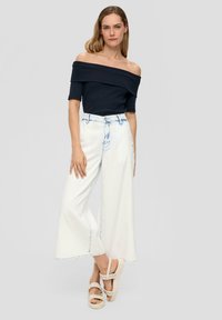 Marinebluse mit schulterfreiem Schnitt, weiße, weit geschnittene Jeans mit hellblauen Akzenten und geflochtenen, beigen Plateau-Sandalen, die vor einem neutralen Hintergrund stehen.