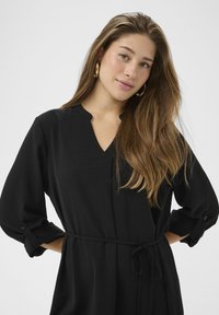 Rochie neagră cu decolteu în V, mâneci lungi, cu cordon în talie și manșete cu nasturi. Confecționată dintr-un material texturat, evidențiind un design simplu și elegant.