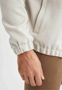 Main masculine reposant à côté du poignet d'une veste beige avec ourlet élastique, portant un pantalon texturé marron.