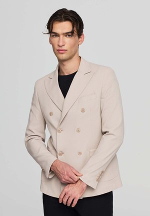 Blazer beige doppiopetto in tessuto testurizzato, con quattro bottoni, rever a punte e una tasca sul petto. Indossato sopra una camicia nera.