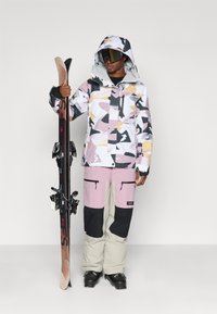 Skiklær som inkluderer en flerfarget jakke med geometrisk mønster i hvitt, rosa, svart og gult, kombinert med lyse rosa og beige snøbukser.