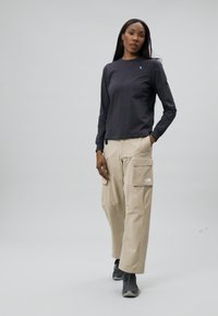 Langarmige donkere top met een klein blauw logo, gecombineerd met lichtbeige cargobroek met meerdere zakken en een logo, aangevuld met zwarte schoenen.