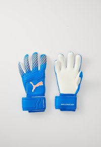 Kék kapuskesztyűk fehér tenyérrel, texturált tapadással, narancssárga kiegészítőkkel és kék pánttal rendelkeznek, amelyen a "4MM ULTRA GRIP" felirat található.