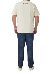 Weißes T-Shirt mit kurzen Ärmeln und rundem Ausschnitt, mit Etikett hinten, kombiniert mit dunkelblauer Jeans und weißen Turnschuhen.