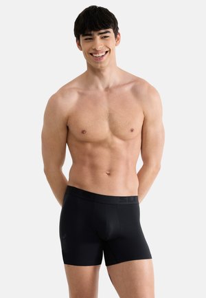 Czarne bokserki o gładkiej fakturze i elastycznym pasku w talii ozdobionym subtelnym wzorem z logo, noszone przez męskiego modela o neutralnym wyrazie twarzy.