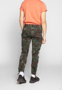 Spodnie cargo w kamuflażowym wzorze w odcieniach zieleni i brązu. Wyposażone w regulowane mankiety i wiele kieszeni. Połączone z krótkim, pomarańczowym t-shirtem.