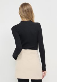Une femme aux longs cheveux châtain clair porte un haut à manches longues noir côtelé et une mini-jupe en velours côtelé beige devant un fond blanc uni.