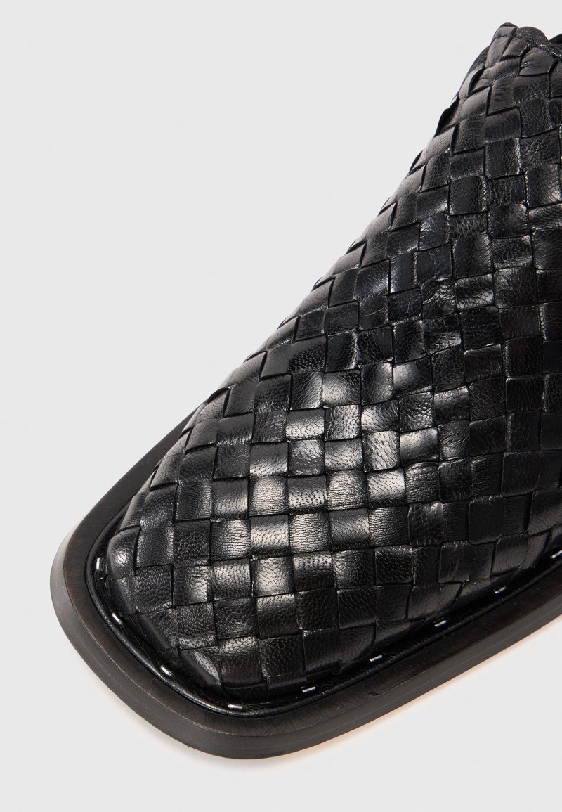 Zwarte leren schoen met vierkante neus, voorzien van een geweven patroon en een zwarte zool met zichtbare stiksels.