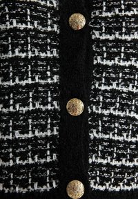 Tissu tweed noir et blanc avec un tissage texturé, panneau central en maille noire et trois boutons ronds dorés avec des motifs complexes.