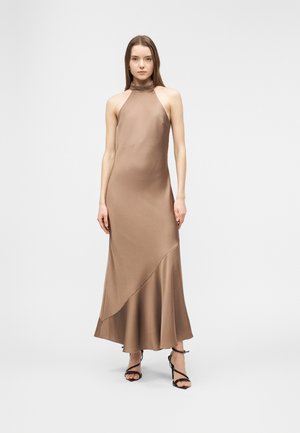 Femme debout portant une robe en satin marron sans manches avec un col halter et un ourlet asymétrique à volants, ainsi que des escarpins noirs à fines lanières.