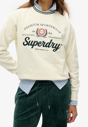 Biała bluza Superdry z emblematem sportowym noszona na niebieską koszulę w paski i zielone spodnie ze sznurkiem na stojącej osobie.