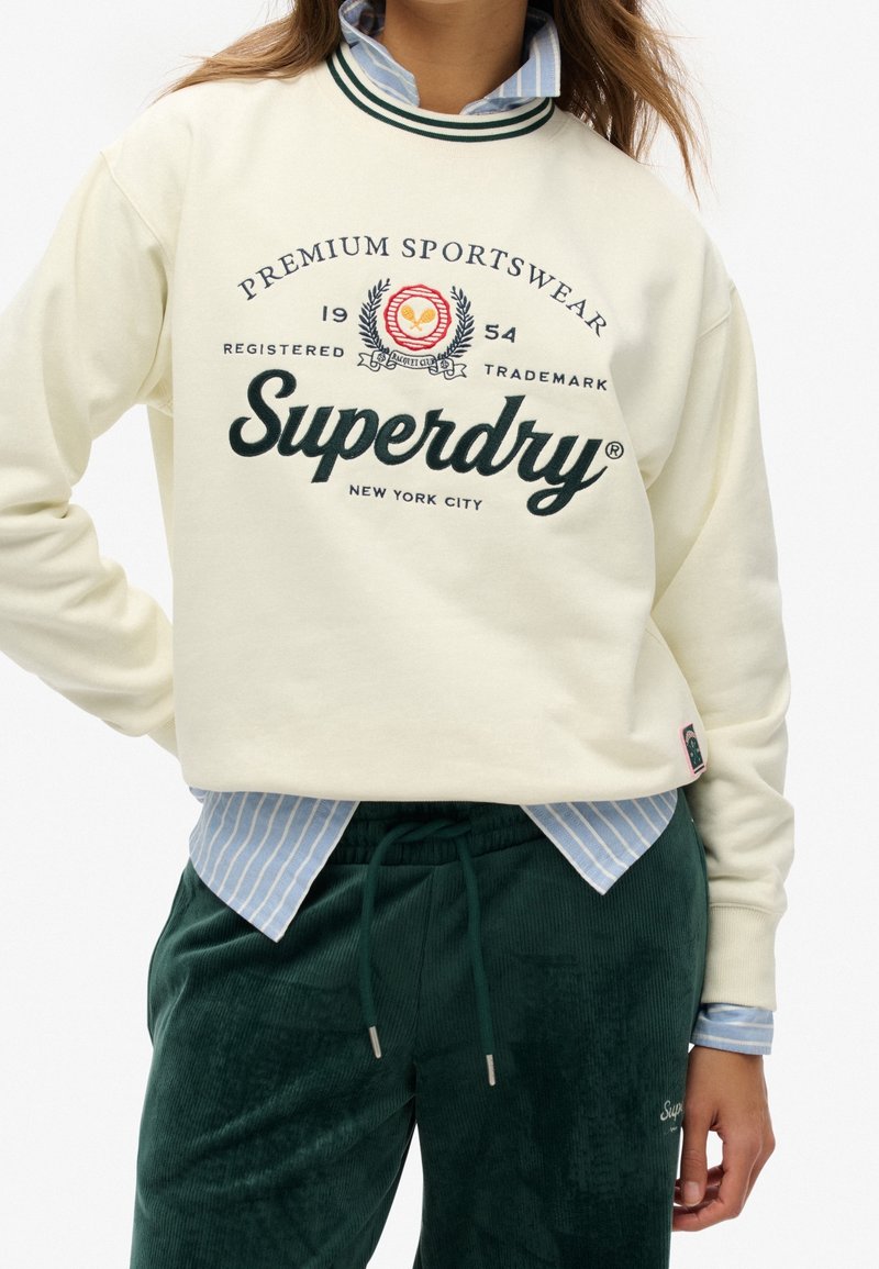 Sweat-shirt blanc Superdry avec emblème sportif porté sur une chemise rayée bleue et un pantalon vert à cordon sur une personne debout.